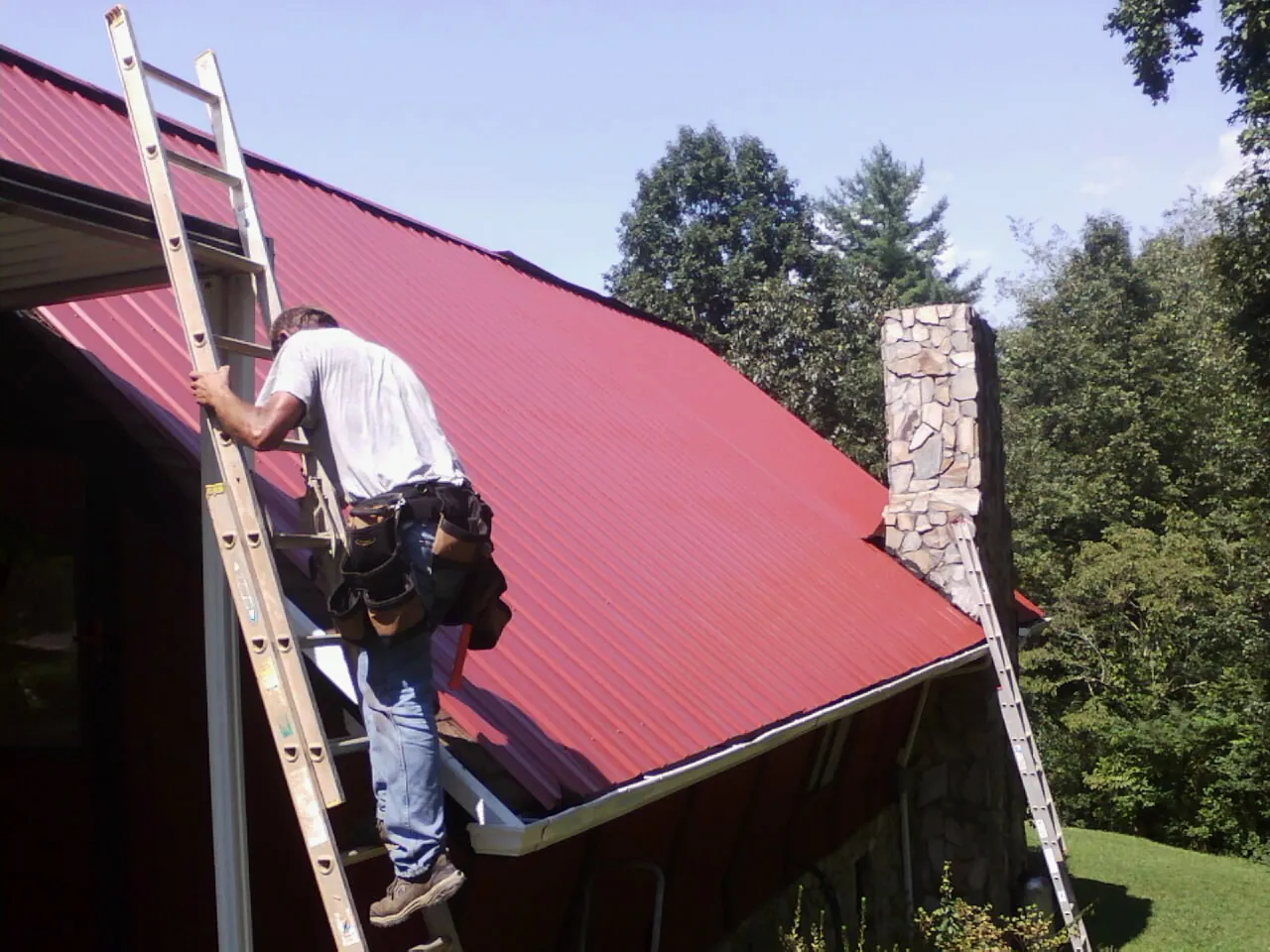 Roofer on a ladder beside a red metal roof and stone chimney — Soffit & Fascia Repair by Roofing Brothers in Deatsville